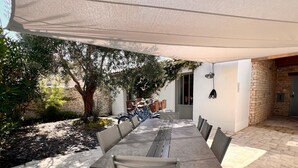 An outdoor dining area with a long table and chairs, shaded by a canopy, surrounded by stone walls and greenery. - House RIVAGE       . (Sainte-Marie-de-Ré)