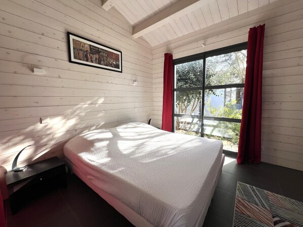 A bedroom with a bed, a window with red curtains, and a framed picture on the wall. - House RIVAGE . (Sainte-Marie-de-Ré)