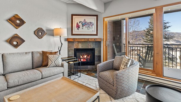 Una sala de estar acogedora con chimenea, un sofá, un sillón estampado y un amplio ventanal con vistas al exterior.