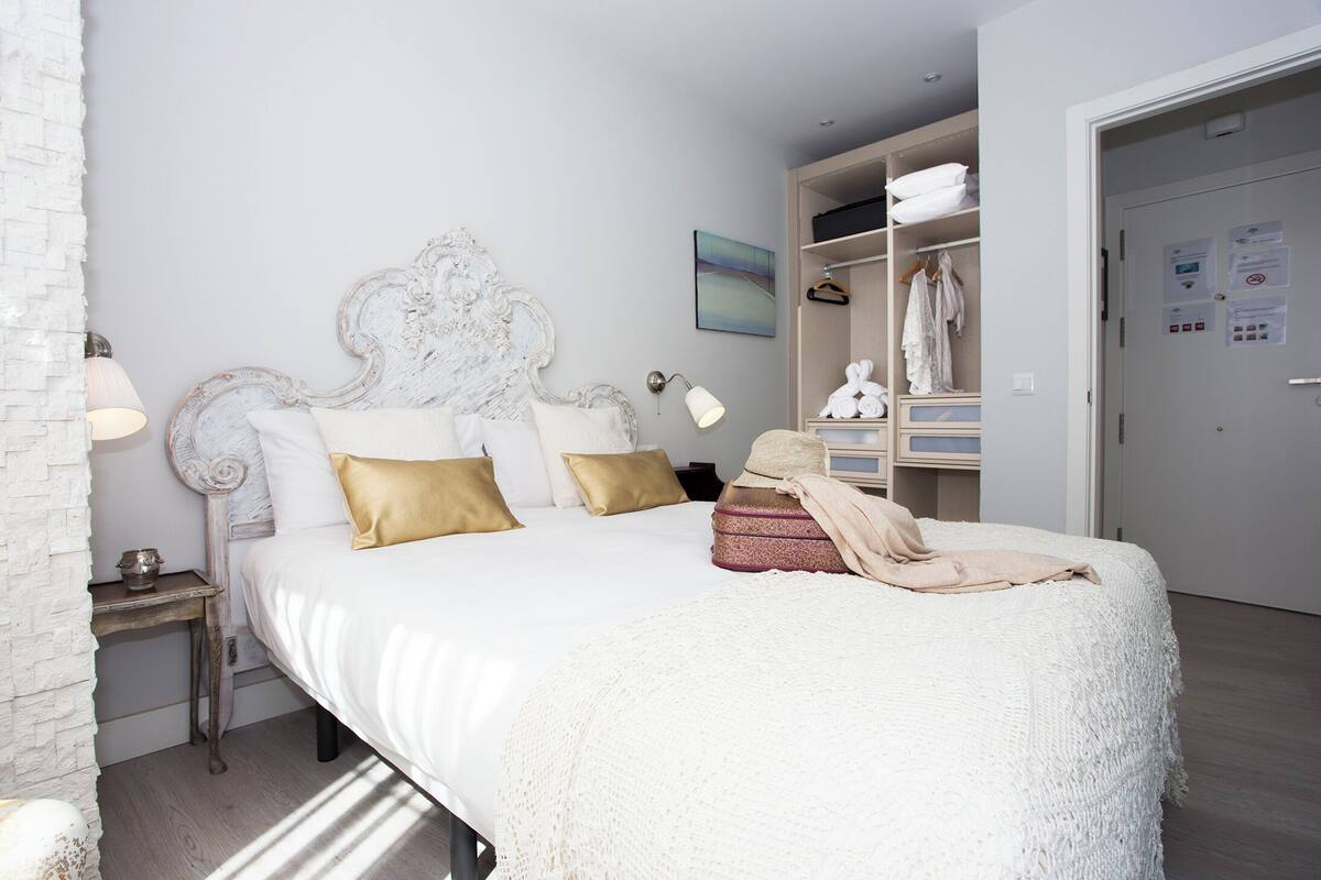 A neatly made bed with a white headboard, a bedside table with a lamp, and a wardrobe with hanging clothes.