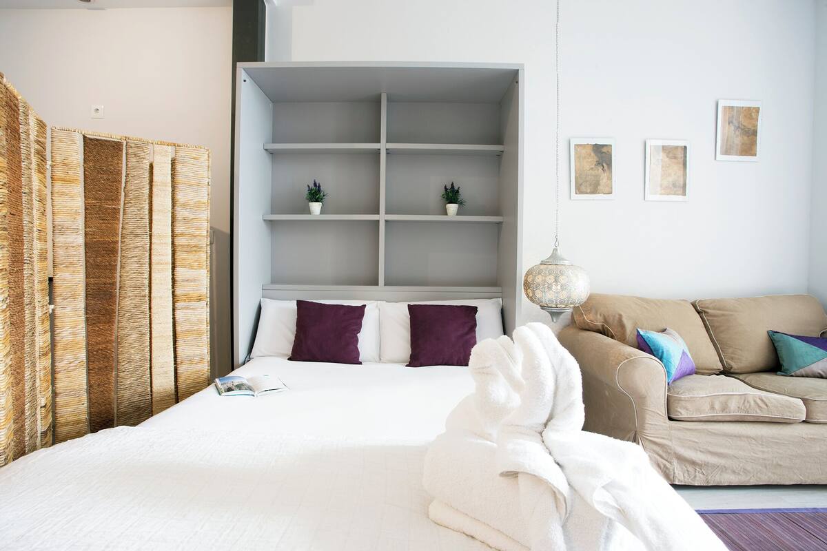 A neatly arranged hotel room with a bed, a sofa, a shelving unit with plants, and artwork on the wall.