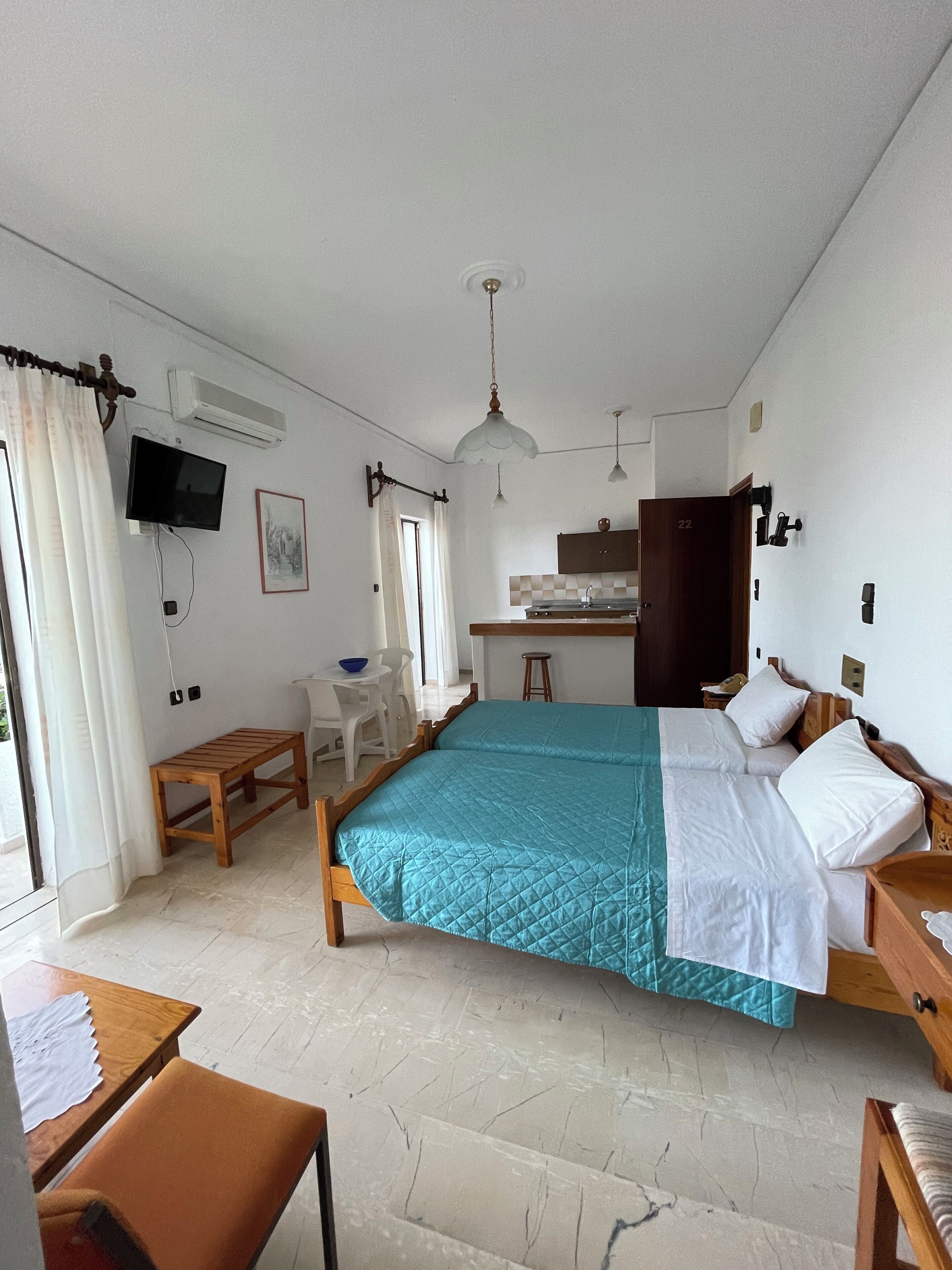 A bedroom with a bed, a desk, a chair, a small table, a TV, and an air conditioning unit.