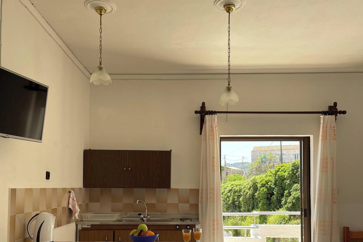 A modern kitchen with a central island, bar stools, a flat-screen TV, and a view of greenery outside.
