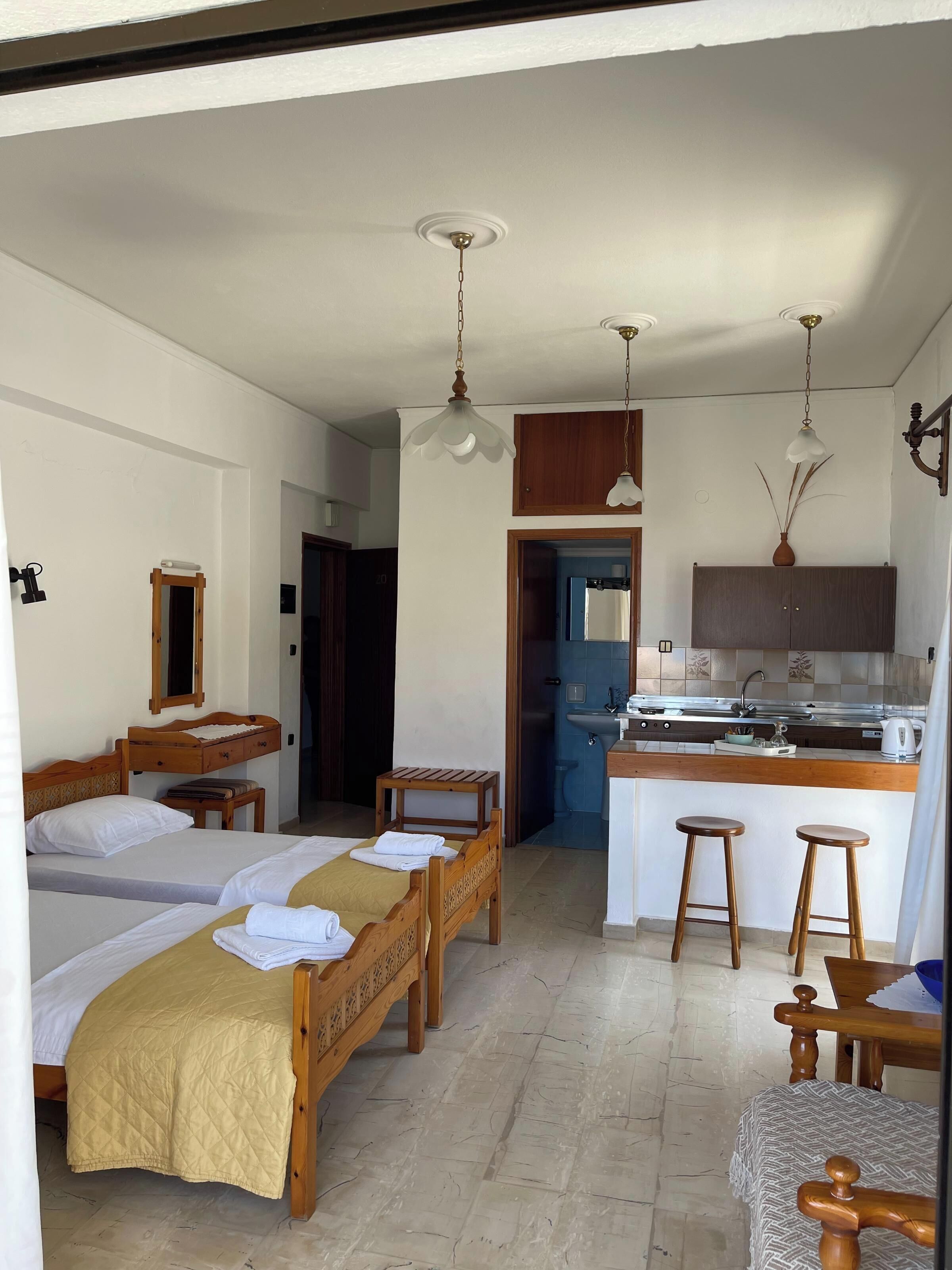A hotel room with two beds, a wooden bench, a small table, a kitchen area with a sink and stools, and a mirror on the wall.