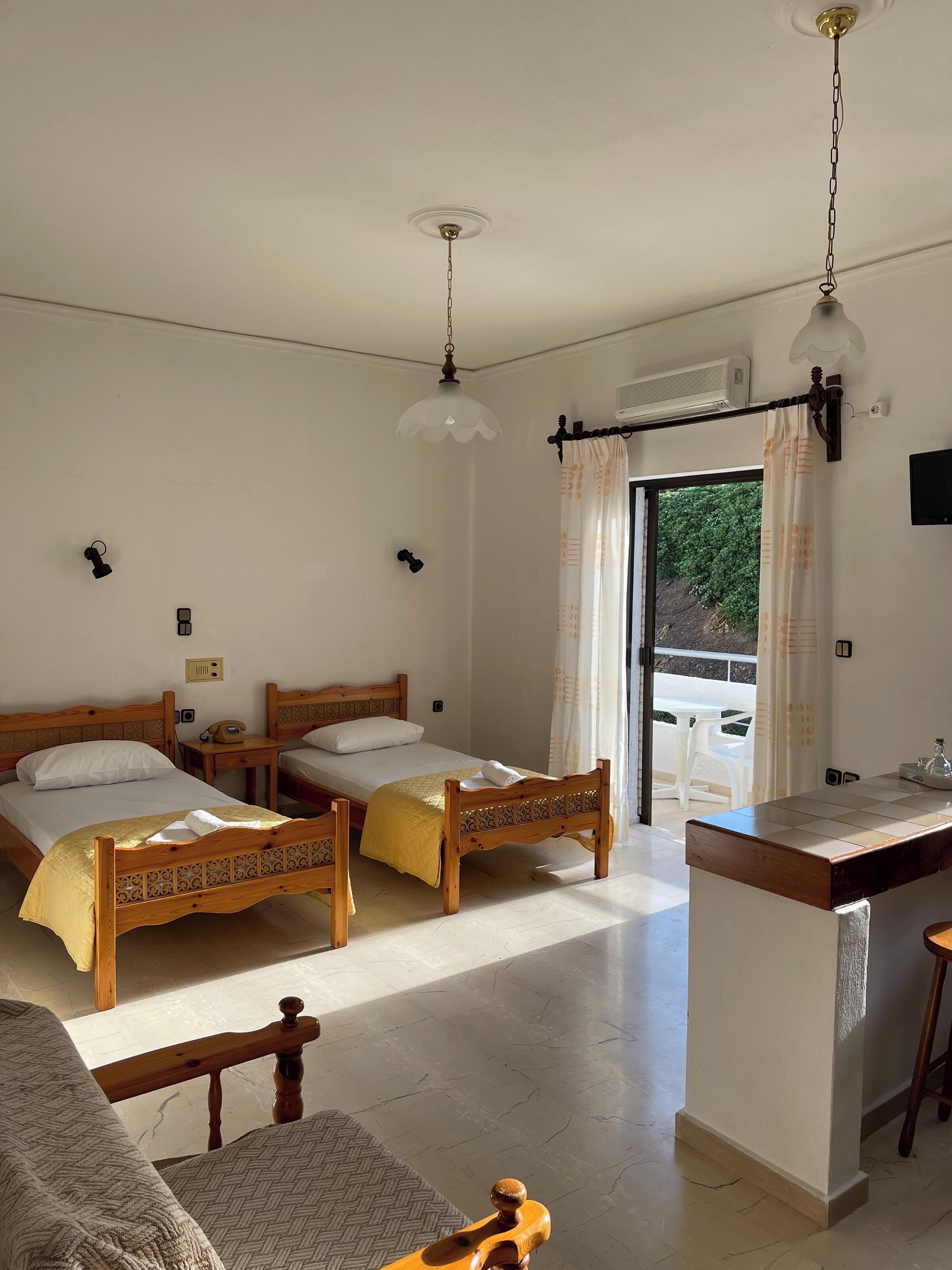 Ein Hotelzimmer mit zwei Holzbett, einem kleinen Tisch, einem Sessel, einer Küchenzeile und Blick ins Grüne.
