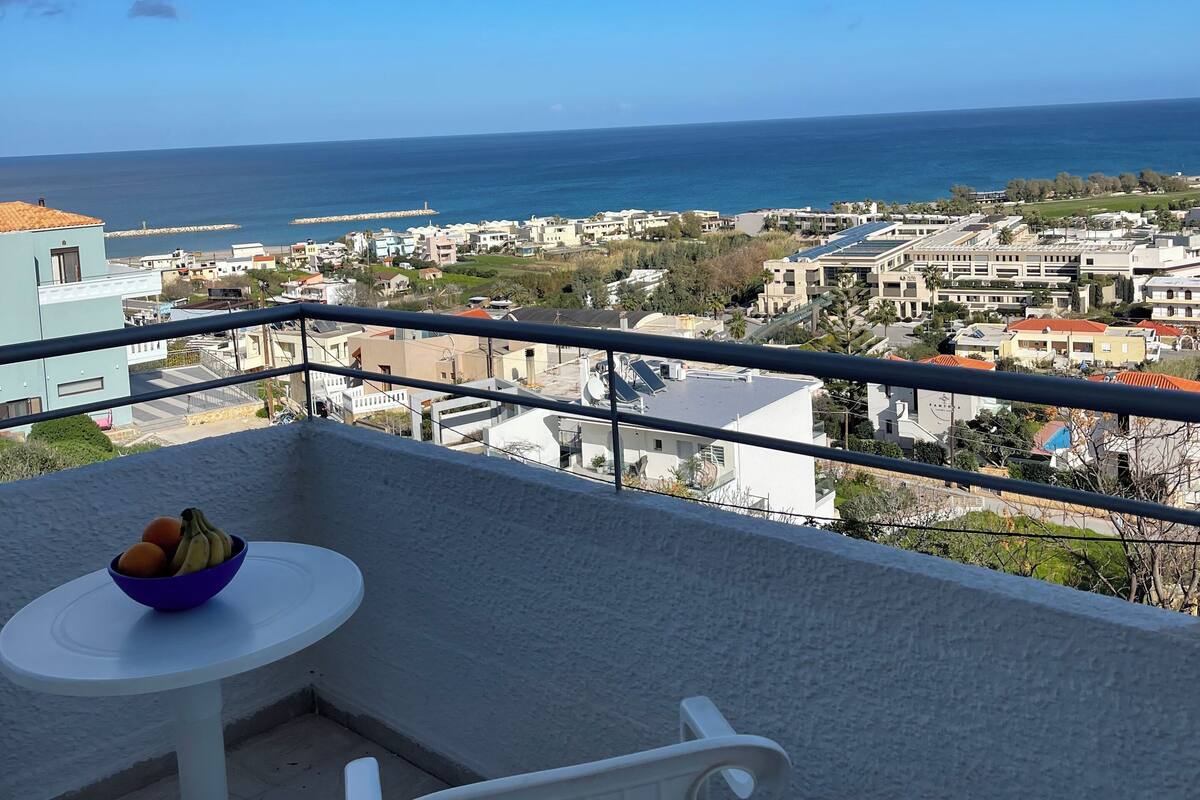A balcony with a table and chairs, overlooking a coastal town and the sea.