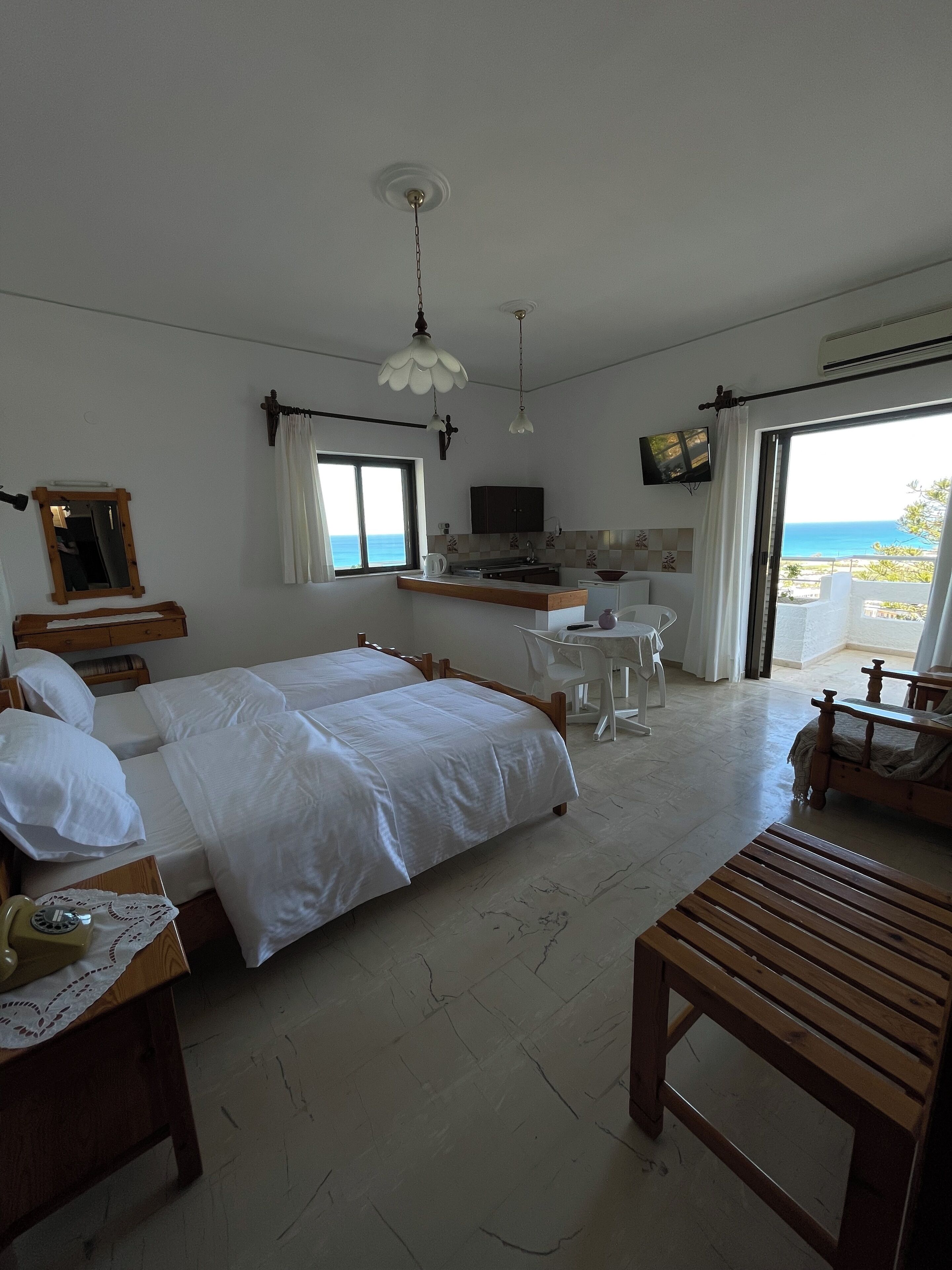 Un dormitorio con cama, mesita de noche y banco de madera. Cuenta con una kitchenette, televisión y un balcón con vista al mar.