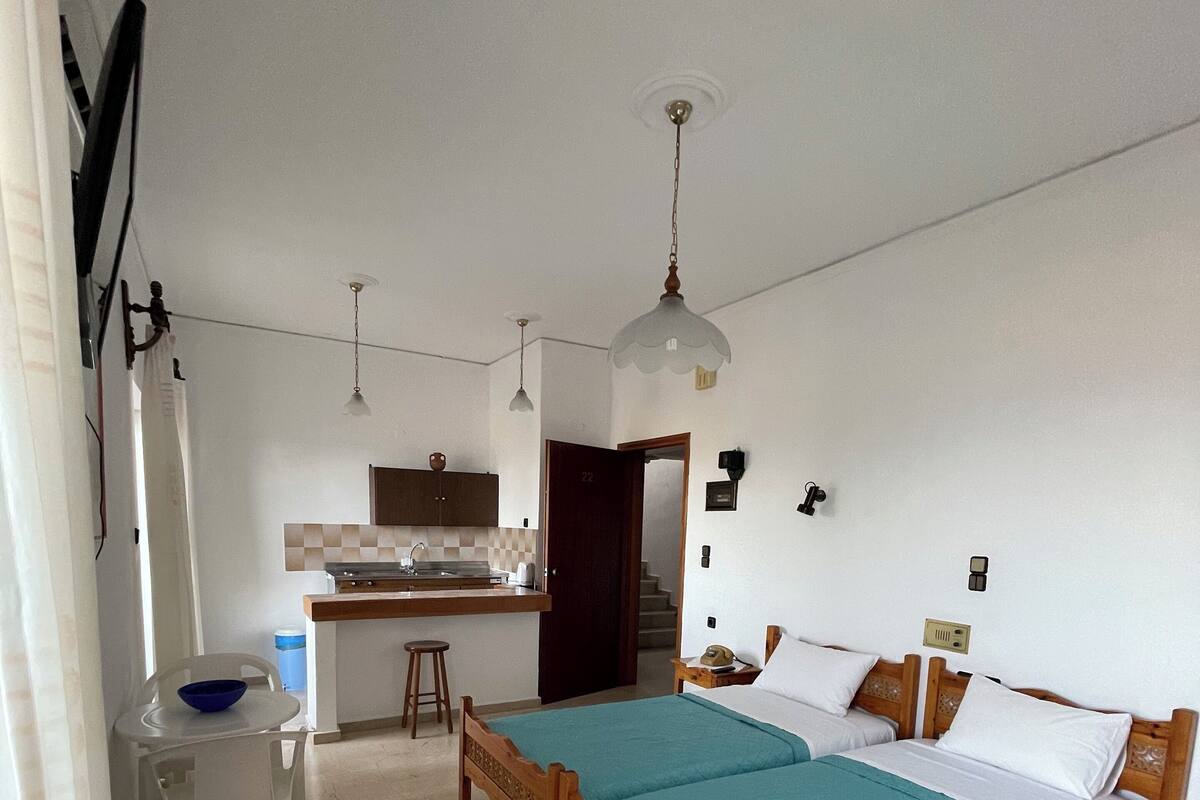 A bedroom with a wooden bed, a small desk, and a kitchenette in the background.