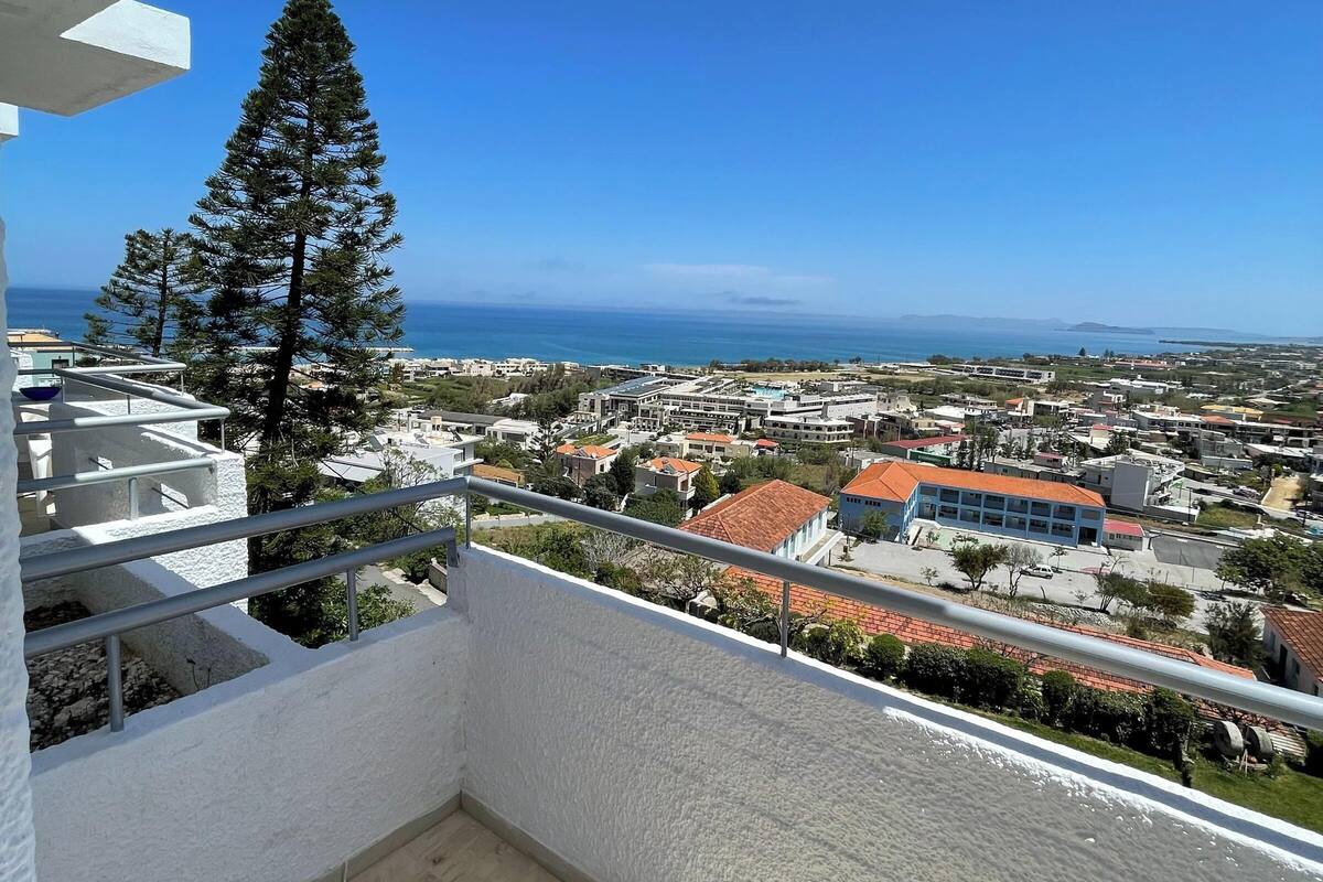 A balcony with a view of a coastal town, a tall pine tree, and a clear blue sky.
