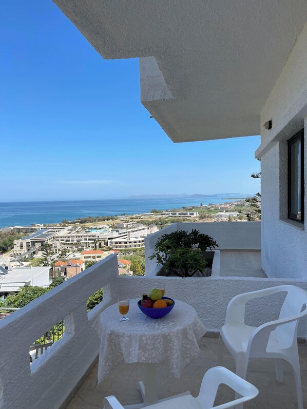 A balcony with a table set for two, a bowl of fruit, and a glass of orange juice. The view includes a body of water and distant buildings. - Aeolos Apartments (Platanias)
