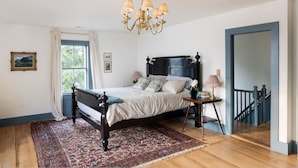 A bedroom with a bed, a chandelier, a nightstand, a window with curtains, a wooden floor, and a staircase.