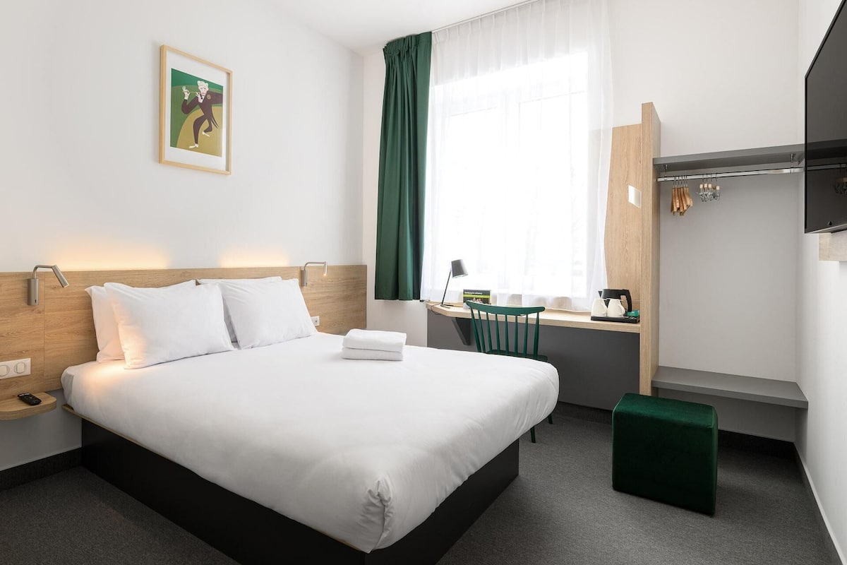 A hotel room with a bed, a desk, a chair, a small stool, a wall-mounted TV, and a painting.