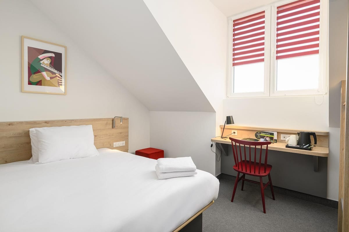 A bedroom with a bed, a desk, a chair, a window with shutters, and a painting on the wall.