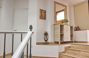 A staircase with a wooden handrail, a white wall with a framed artwork, a wooden cabinet with drawers, and a mirror reflecting a room.