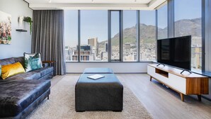 Una sala de estar moderna con un ventanal grande, un sofá, un reposapiés y un televisor sobre un mueble de madera.