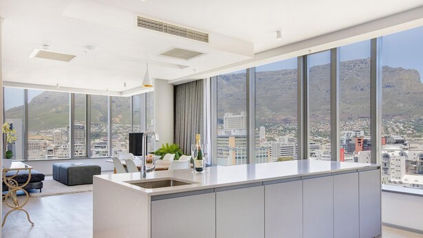 Una cocina moderna con una isla grande, un comedor con mesa y sillas, y vistas a un paisaje urbano a través de ventanas amplias.
