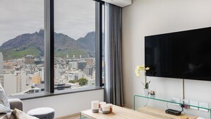 Habitación de hotel moderna con un amplio ventanal que ofrece vistas a la ciudad, un televisor de pantalla plana, un escritorio de madera y una zona de estar acogedora.