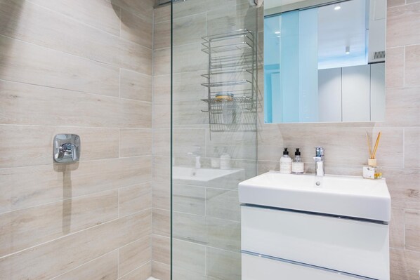 Un baño moderno con cabina de ducha de vidrio, un lavamanos blanco con espejo y un estante de vidrio con artículos de aseo.
