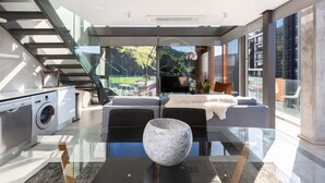 Una sala de estar moderna con un sofá, una mesa de centro y un televisor. Se ve una escalera con barandas de vidrio que conduce a un espacio al aire libre.