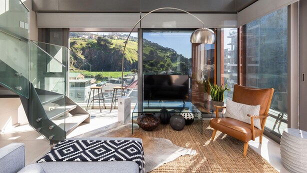 Una sala de estar moderna con una escalera de cristal, un sofá de cuero, una mesa de centro y vistas a una ladera verde.