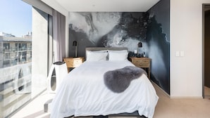 Un dormitorio moderno con una cama grande, dos mesitas de noche, vistas a la ciudad y un mural único en la pared.