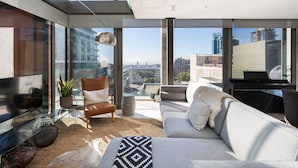 Una sala de estar moderna con sofá, sillón y televisor, ventanas amplias con vistas a la ciudad y una mesa de vidrio con objetos decorativos.