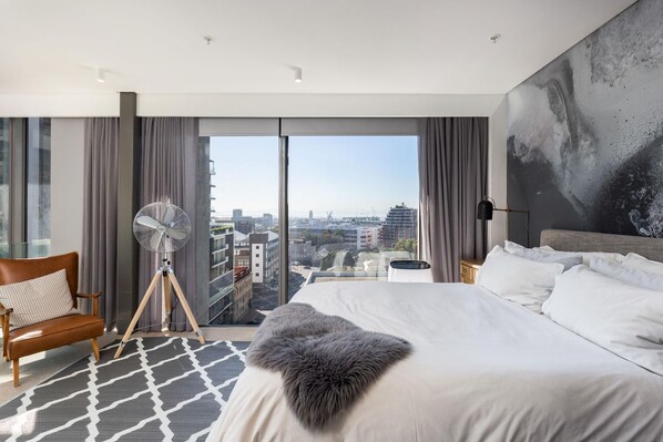 A modern hotel room with a large bed, a view of the city, a standing fan, and a large window.
