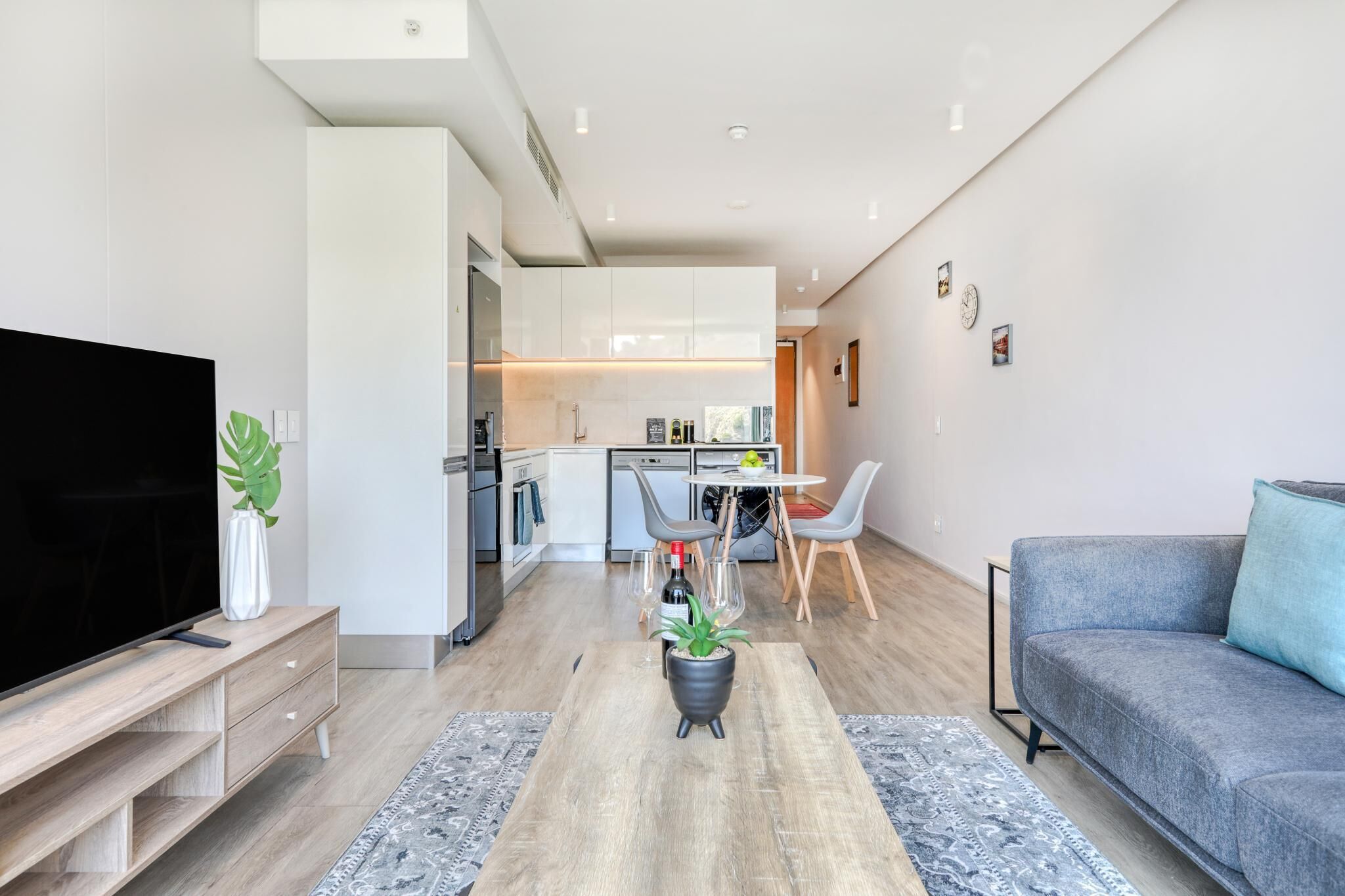 A modern living room with a flat-screen TV, a grey sofa, a wooden coffee table with a plant, and a dining area with a table and chairs.