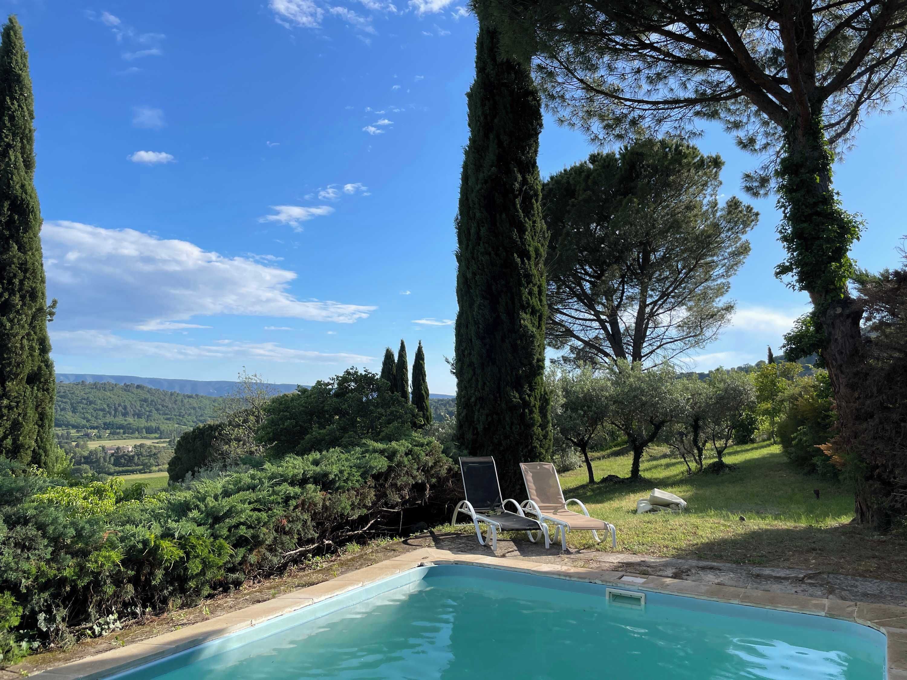 A swimming pool with lounge chairs, surrounded by tall trees and a view of a hilly landscape.