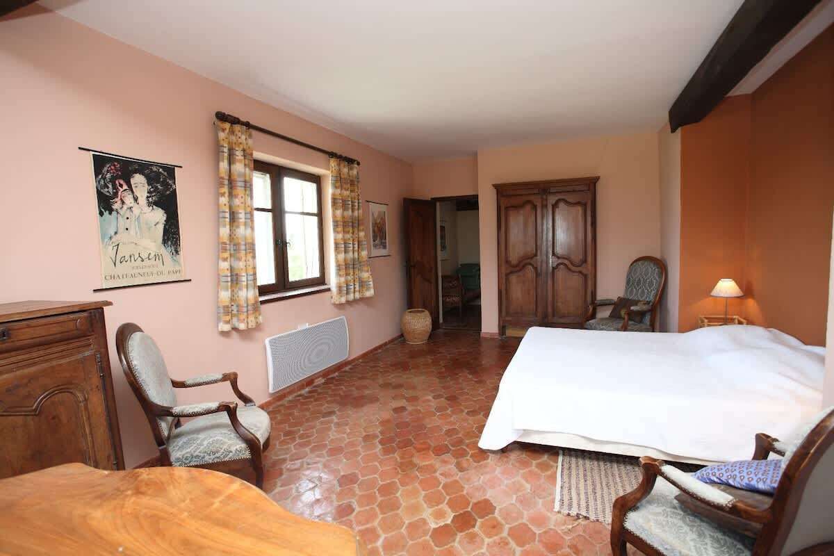 A bedroom with a bed, two chairs, a wooden dresser, a window with curtains, a poster on the wall, and a door leading to another room.
