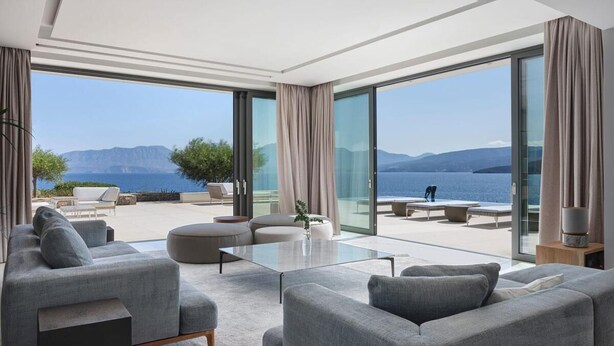 Ein modernes Wohnzimmer mit einem großen Glasfenster, das auf einen Balkon mit Blick auf das Meer und die Berge führt.