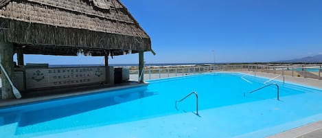 A swimming pool with a thatched-roof structure, clear blue sky, and a distant view of the beach.