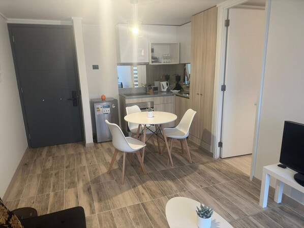 Un espace de vie compact comprenant un coin cuisine, un coin repas avec une table ronde et des chaises, et un téléviseur posé sur un meuble.