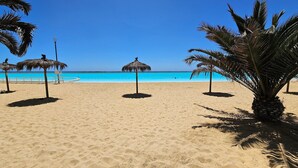 A beach with clear blue water, palm trees, and thatched umbrellas. - Oceana Suites Laguna del Mar (La Serena)