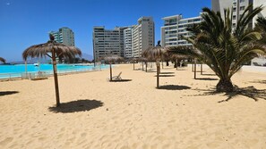 A beach with palm trees, a pool, and high-rise buildings in the background.