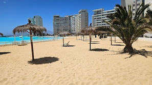 Una playa con palmeras, una piscina y edificios altos al fondo.