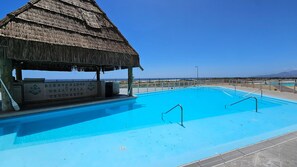 Une piscine dotée d’une structure surmontée d’un toit de chaume, un ciel bleu azur et une vue lointaine sur la plage.