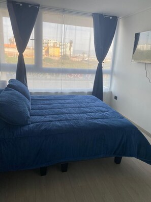 Un dormitorio con cama, cortinas, ventana y televisor montado en la pared.