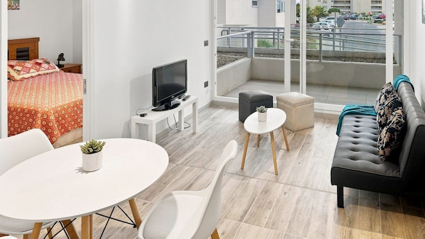 Ein modernes Hotelzimmer mit Balkon, einem Bett mit gemustertem Überwurf, einem Fernseher auf einem Standfuß, einem kleinen Tisch mit Pflanze und einem Sofa mit Kissen.
