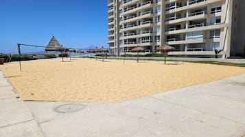 A sandy beach area with a volleyball net, umbrellas, and a building in the background.