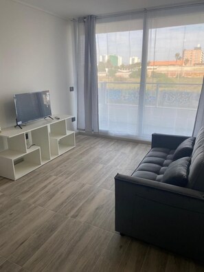 Een moderne woonkamer met een grijze bank, een flatscreen televisie op een witte kast, en een groot raam met uitzicht op gebouwen en groen.