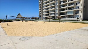 A sandy beach area with a volleyball net, umbrellas, and a building in the background. - Oceana Suites Laguna del Mar (La Serena)