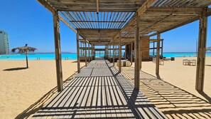 Houten terras dat uitkomt op een strand met helderblauw water en een gebouw in de verte.