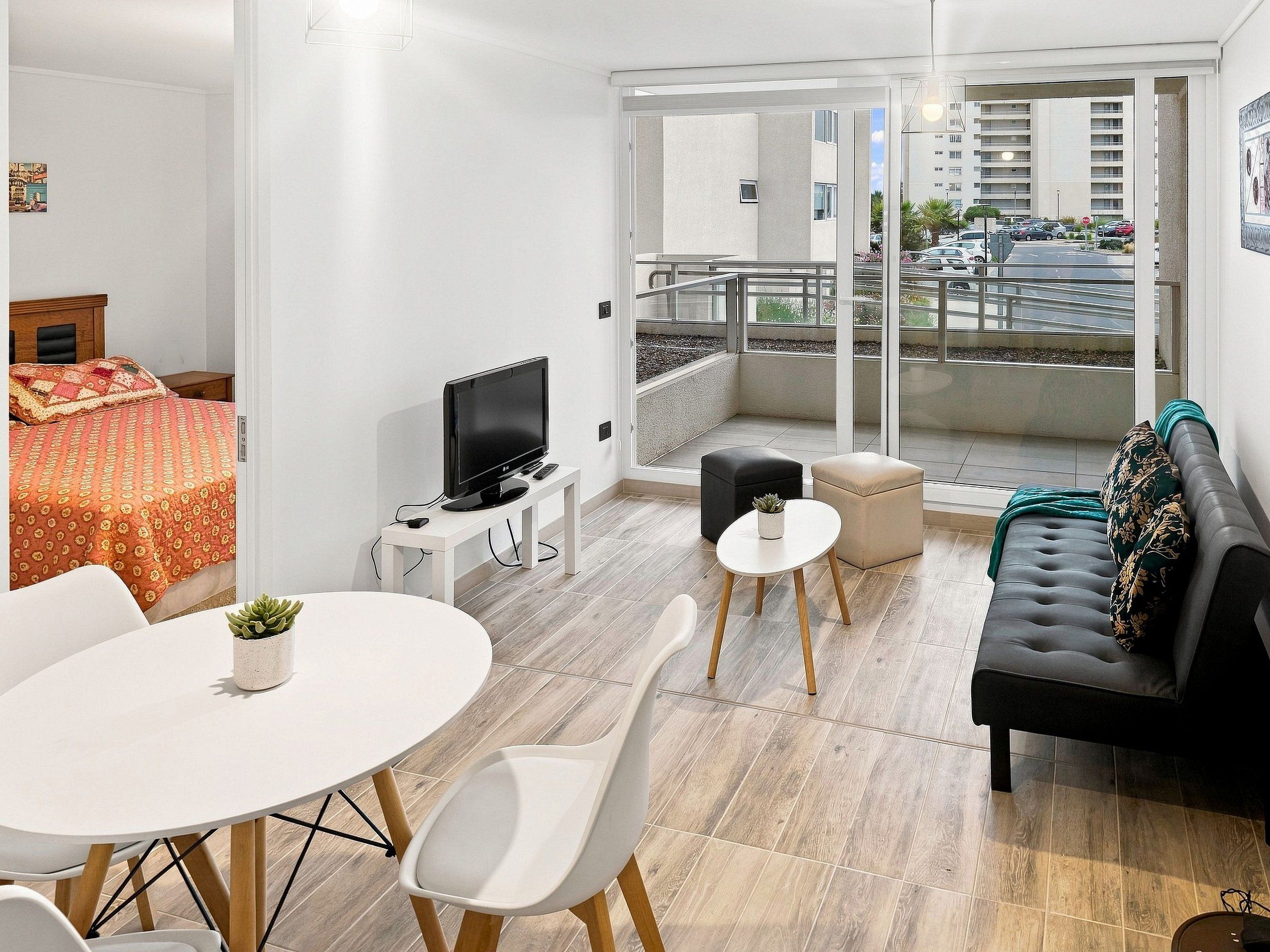 A modern living room with a sofa, a TV, a dining table, and a balcony with a view of the street.