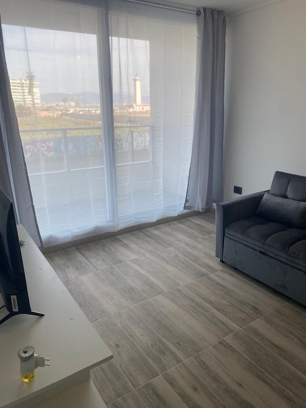 Una habitación con sofá gris, una mesa blanca con toma de corriente y un ventanal con cortinas traslúcidas.