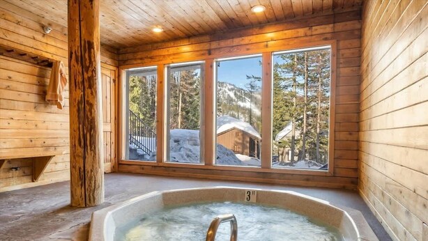 A wooden cabin with a hot tub, large windows, and a view of snow-covered mountains.