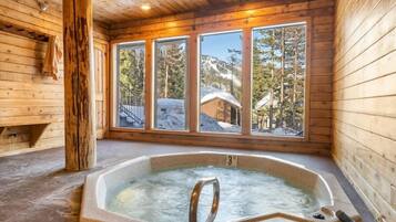 A wooden cabin with a hot tub, large windows, and a view of snow-covered mountains.
