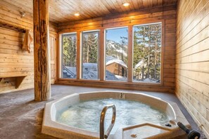 Uma cabana de madeira com hidromassagem, grandes janelas e vista para montanhas cobertas de neve.