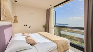 A modern bedroom with a large window, a bed with white linens, a beige throw blanket, and a hanging light fixture. - Hedera Estate, Villa Hedera XX (Dubrovnik)