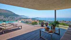 A modern outdoor terrace with a view of a coastal town, a pool, and a table with food and drinks. - Hedera Estate, Villa Hedera XX (Dubrovnik)